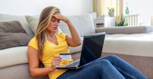 A woman sitting on the floor holding a credit card and looking stressed while using a laptop, representing the financial and emotional impact of identity theft.
